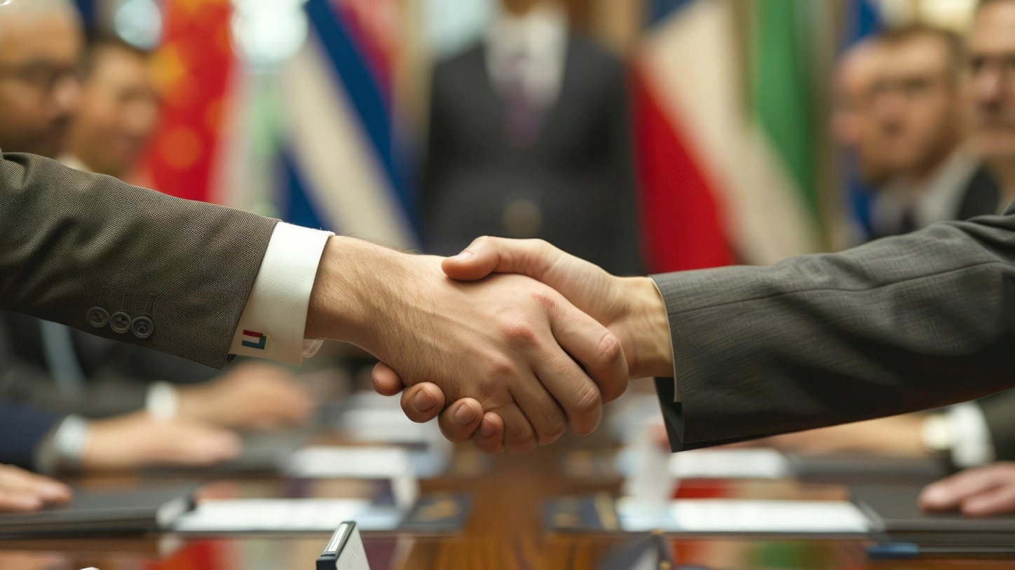 Una stretta di mano tra due uomini d’affari a un tavolo di conferenza, con bandiere internazionali sullo sfondo