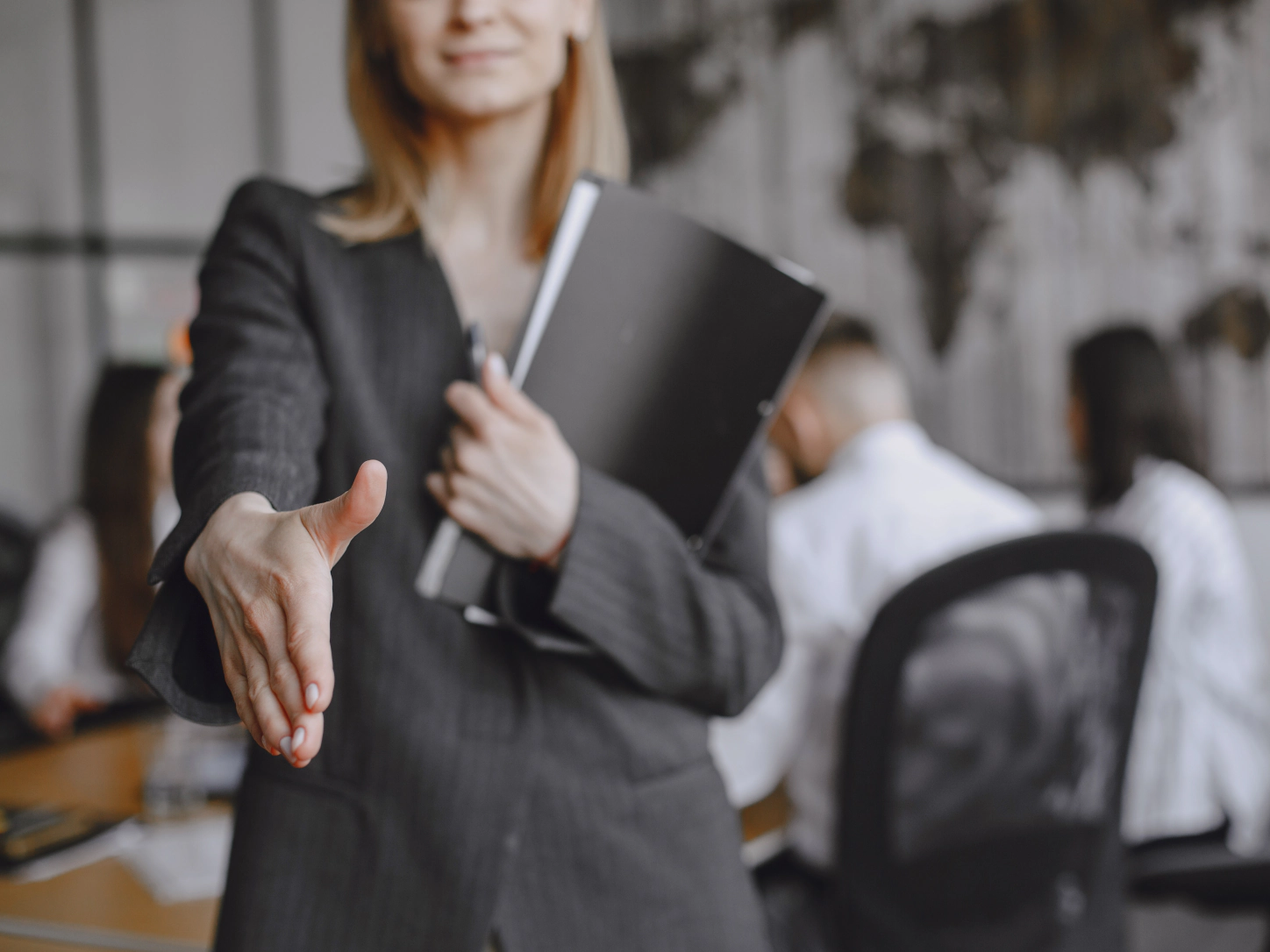 Una professionista con una cartella sotto il braccio tende la mano per salutare, in un ufficio con colleghi al lavoro